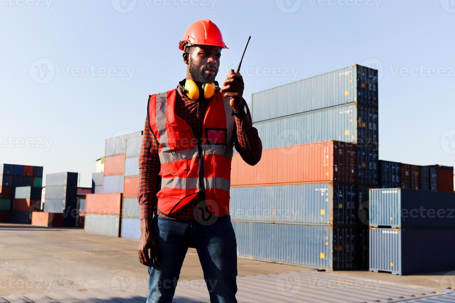 porträtt av ung afroamerikansk ingenjör arbetare man bär säkerhetsljus neonröd färg väst och hjälm, pratar med en kollega med walkies-talkie på logistisk frakt lastcontainergård. foto