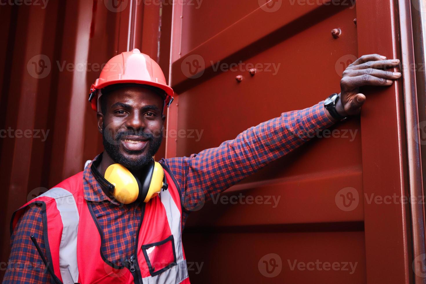 porträtt av glada leende afrikansk amerikansk ung ingenjör arbetare man bär säkerhetsljus neonröd färg väst och hjälm, öppnar en containerdörr på logistisk lastcontainergård. foto