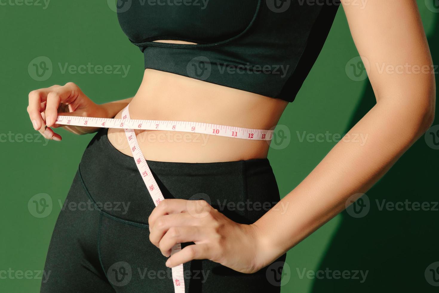 beskära närbild kroppen av sportig kvinna som mäter hennes tunna midja med ett måttband isolerad på grön bakgrund. dynamisk rörelse. styrka och motivation. foto