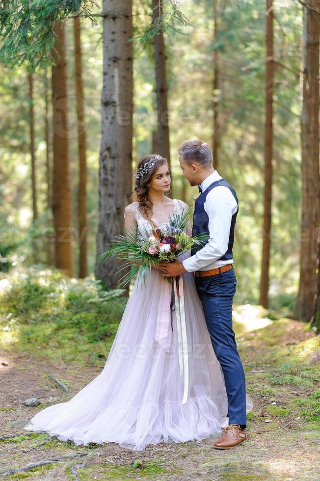 ett attraktivt nygift par, en lycklig och glädjefull stund. en man och en kvinna rakar sig och kysser sig i semesterkläder. bröllopscermonia i bohemisk stil i skogen i frisk luft. foto