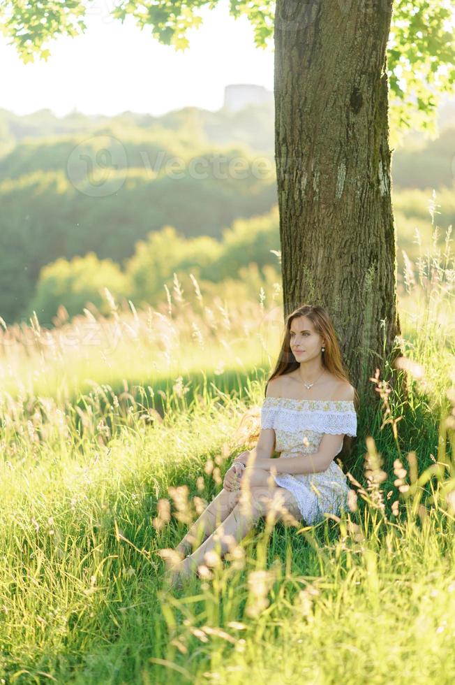 porträtt av en ung vacker flicka i en solklänning. sommarfotosession i parken vid solnedgången. en flicka sitter under ett träd i skuggan. foto