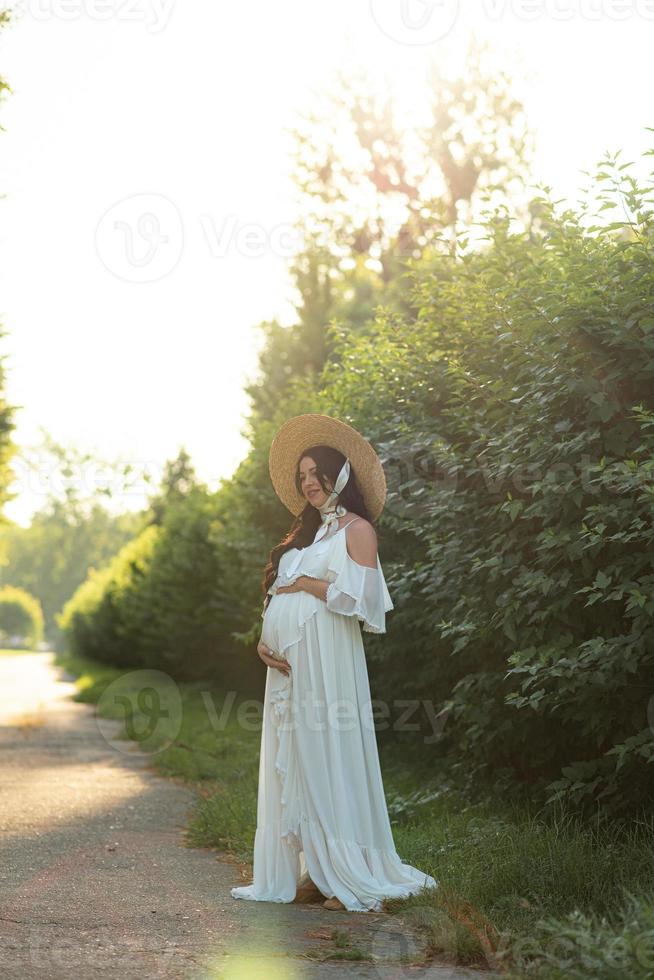 gravid kvinna i en hatt poserar i en klänning på en bakgrund av gröna träd. foto