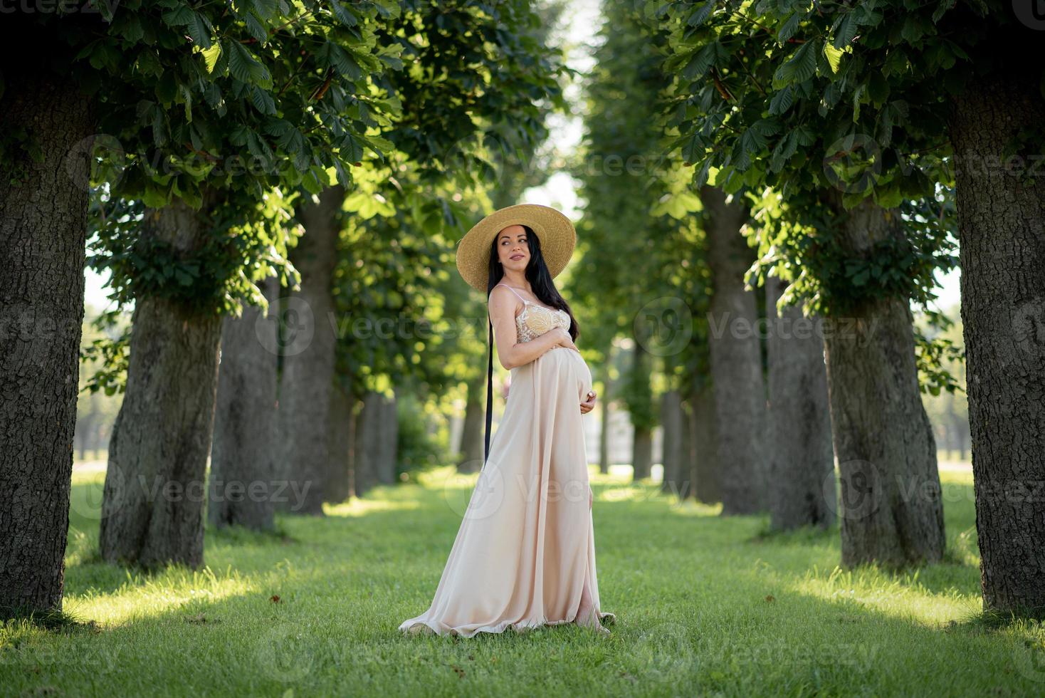 gravid kvinna i en hatt poserar i en klänning på en bakgrund av gröna träd. foto