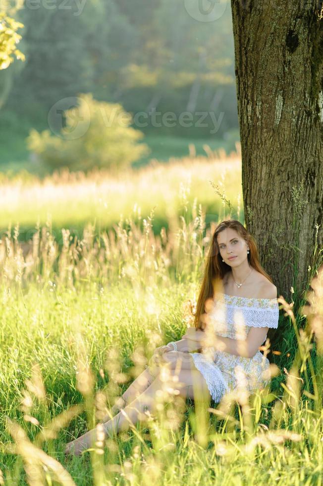 porträtt av en ung vacker flicka i en solklänning. sommarfotosession i parken vid solnedgången. en flicka sitter under ett träd i skuggan. foto