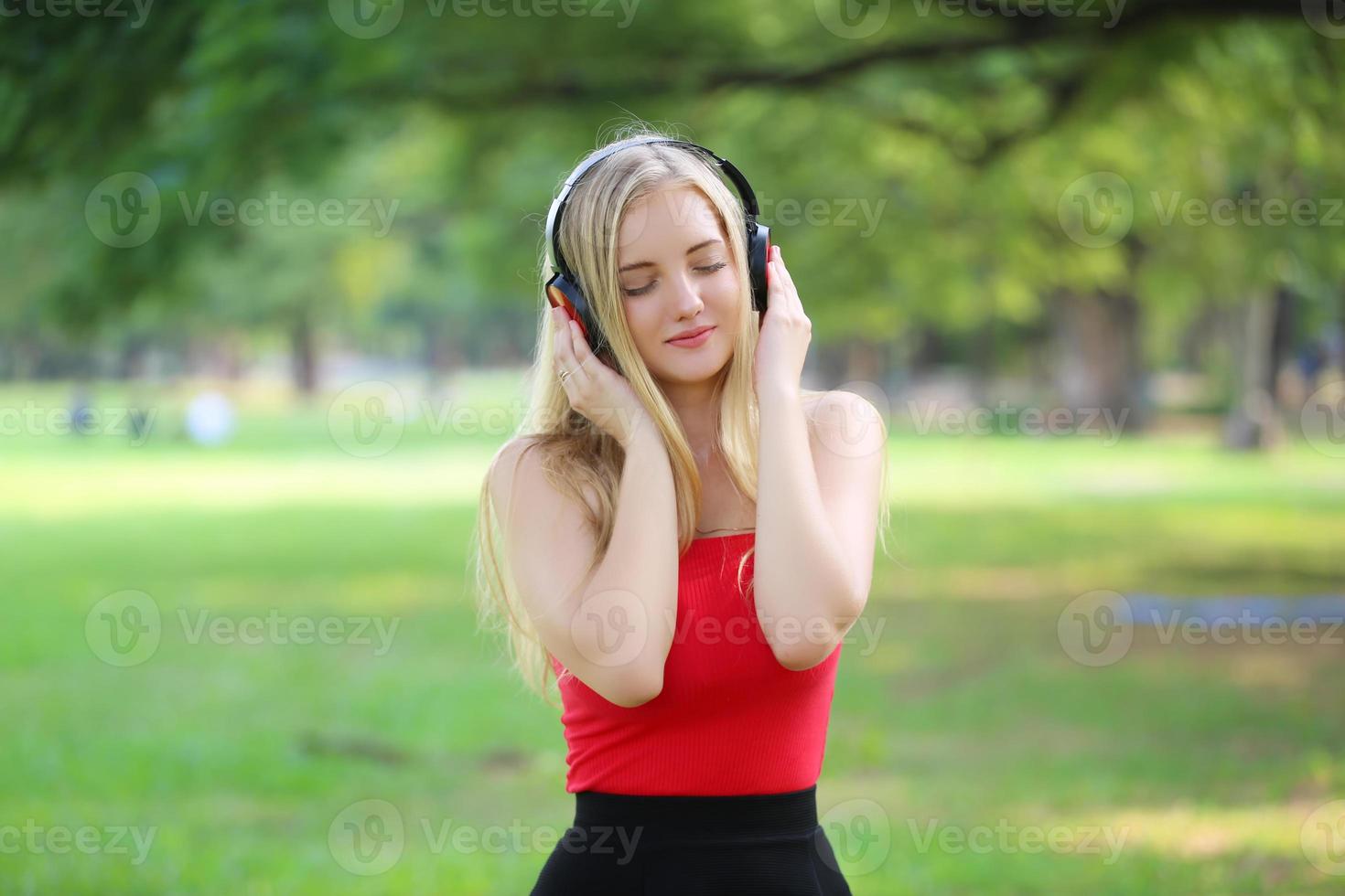 vacker blond kvinna står medan du lyssnar på musik i hörlurar i parken. foto