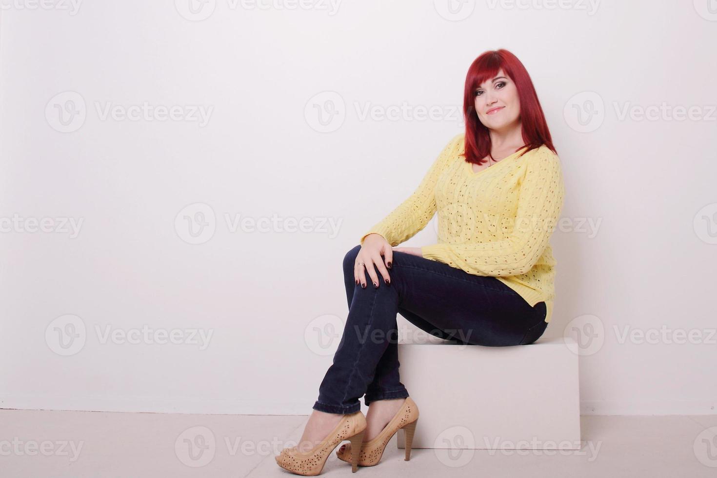 rött hår kvinna i jeans som sitter på vit kub, studiofotografering, kopieringsutrymme foto