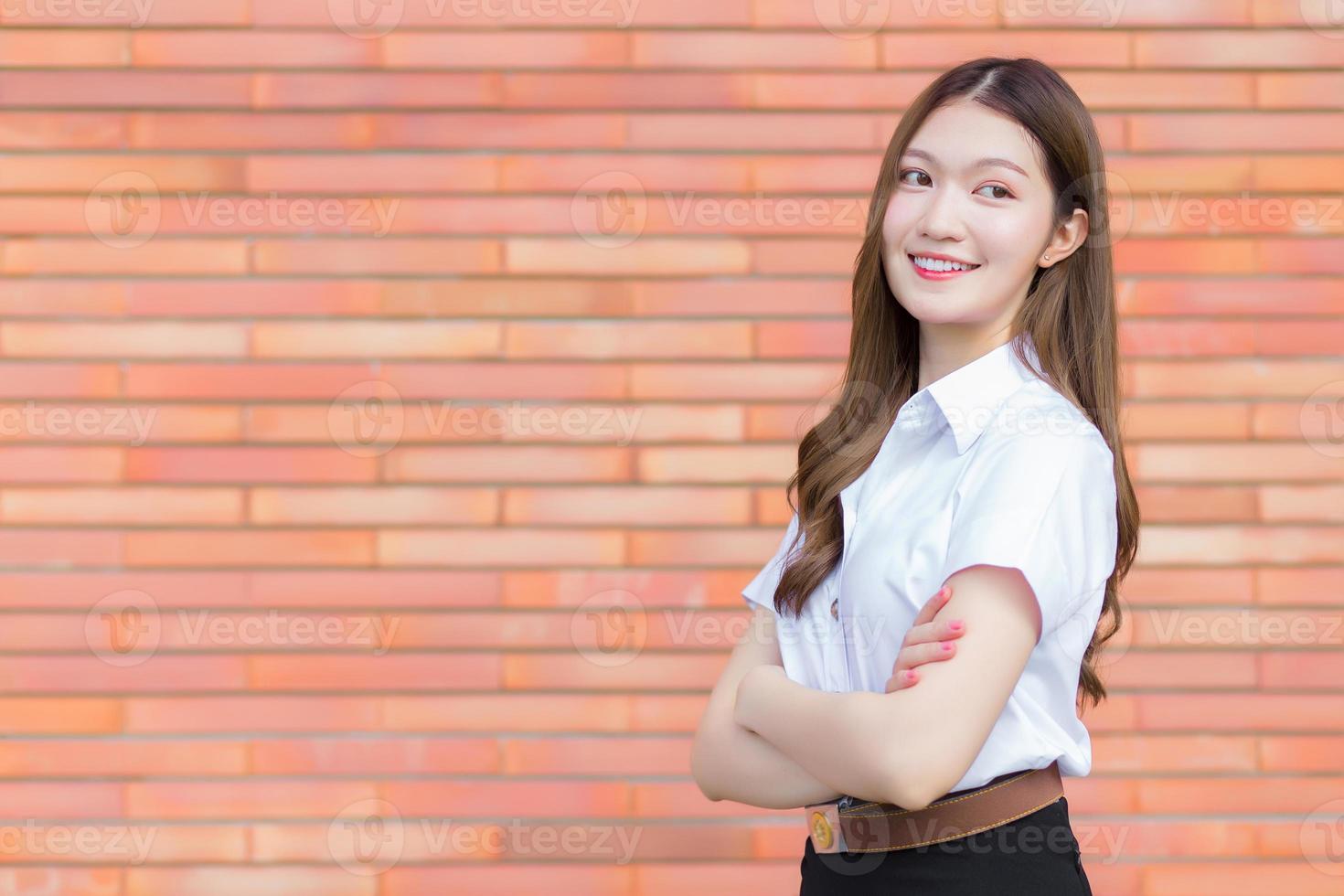 porträtt av en vuxen thai student i universitetsstudentuniform. asiatisk vacker flicka står med armarna i kors på en tegelbakgrund. foto