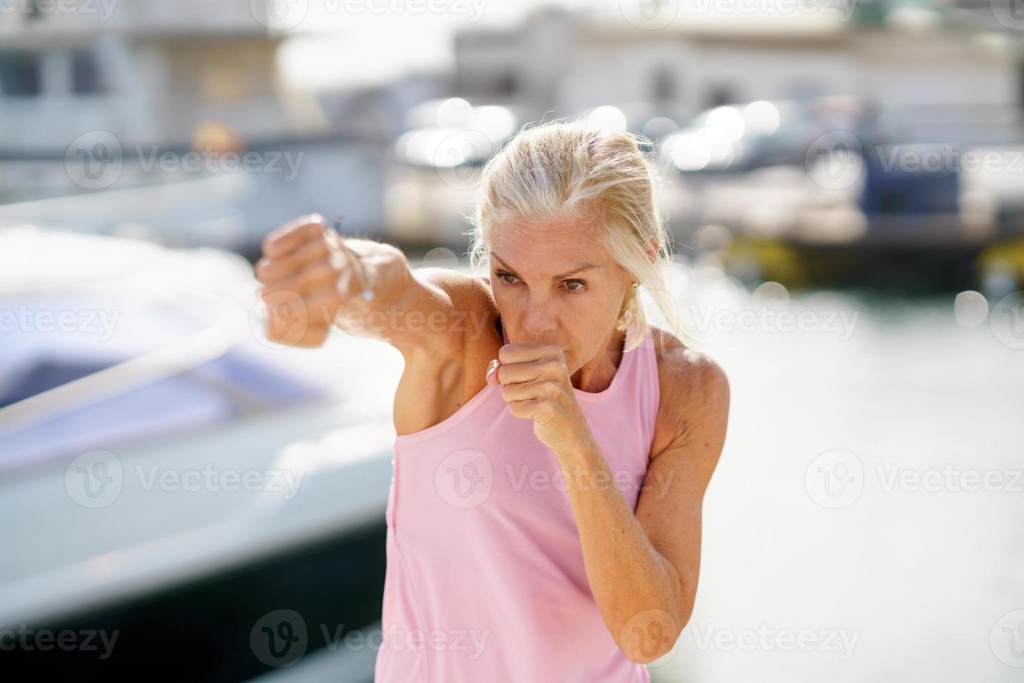 äldre kvinna gör skuggboxning utomhus. senior kvinna gör sport i en kust hamn foto