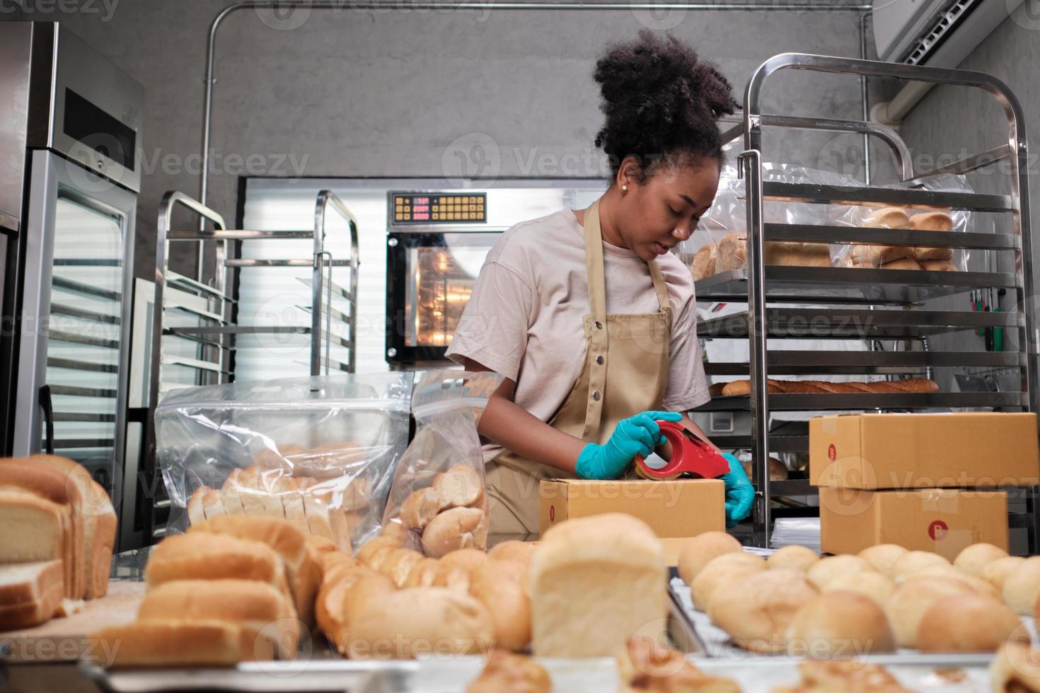 bageri startup leverans av småföretag. en afroamerikansk kvinnlig kock packar handgjorda och nybakade bröd och bakverk i lådor och skickar till kundköp online i kulinariska kök. foto