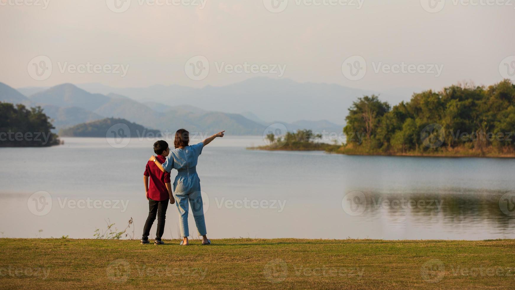 mor och son står bredvid den stora sjön och ser bergsutsikten i bakgrunden, mamma pekar finger mot skogen. idé för familjen turistresor tillsammans till utomhusresan foto