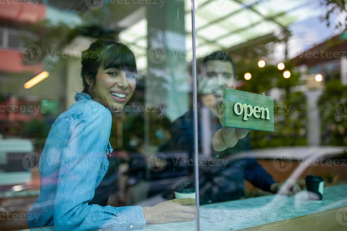 porträtt av leende ägare på hennes café med öppen skylt. kaffeägare i kafé för att välkomna kunden och öppna kaféet på morgonen. foto