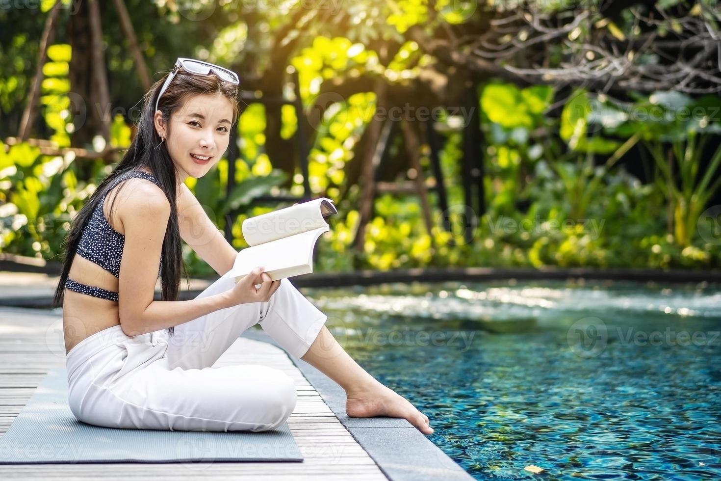 vacker ung asiatisk kvinna sitter på poolkanten utomhus. hon läser bok medan hon kopplar av nära poolen foto