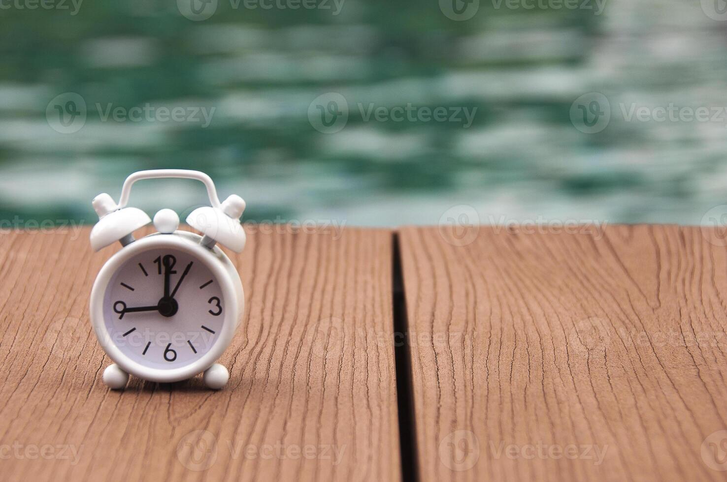 väckarklocka på trägolv med suddig poolbakgrund. klockan ställd på 9.00. morgon koncept. foto