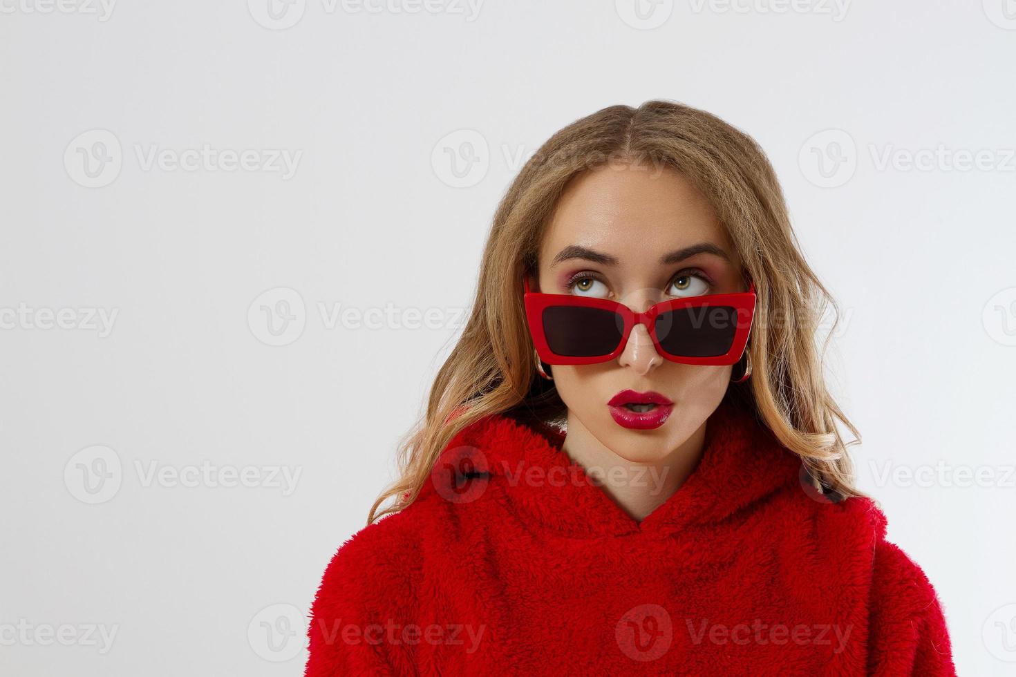 närbild kvinna ansikte med röda läppar, solglasögon hoodie isolerad på vit bakgrund. moderiktig tjej i snygg outfit. makeup, skönhet koncept. vinter höstkläder. chock upphetsat kvinnligt ansikte. närbild foto