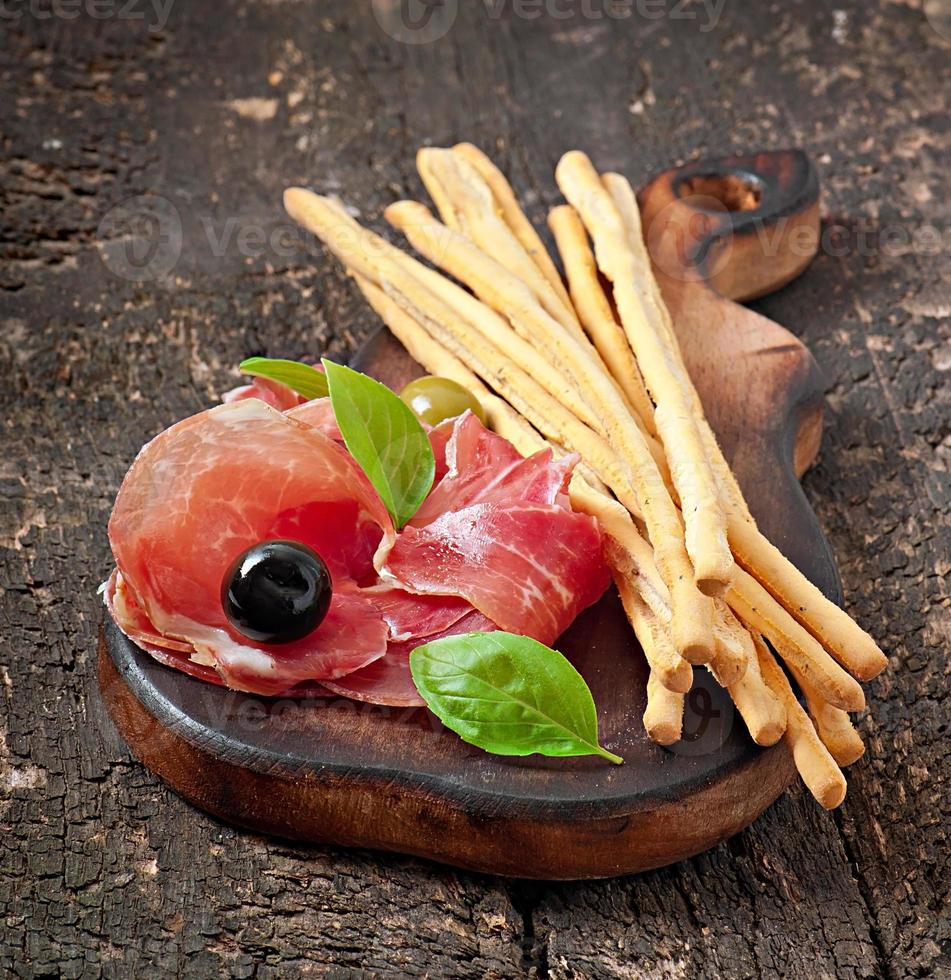 grissini bröd pinnar med skinka, oliver, basilika på gammal trä bakgrund foto