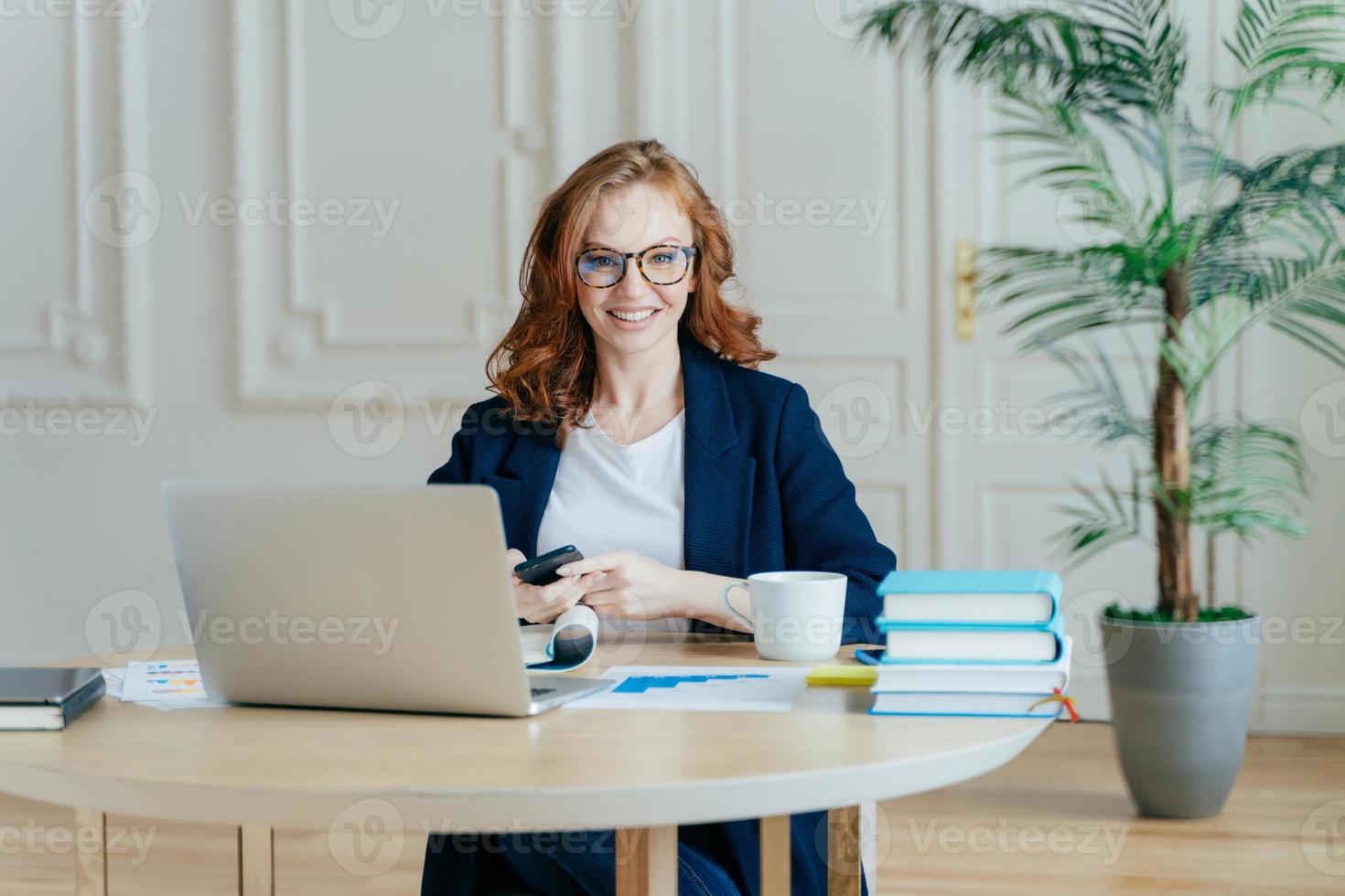 inomhusbild av rödhårig kvinnlig ekonom skickar textmeddelande till kollega, håller en modern smart telefon, poserar på arbetsplatsen, arbetar med finansiella dokument, omgiven av modern teknik foto