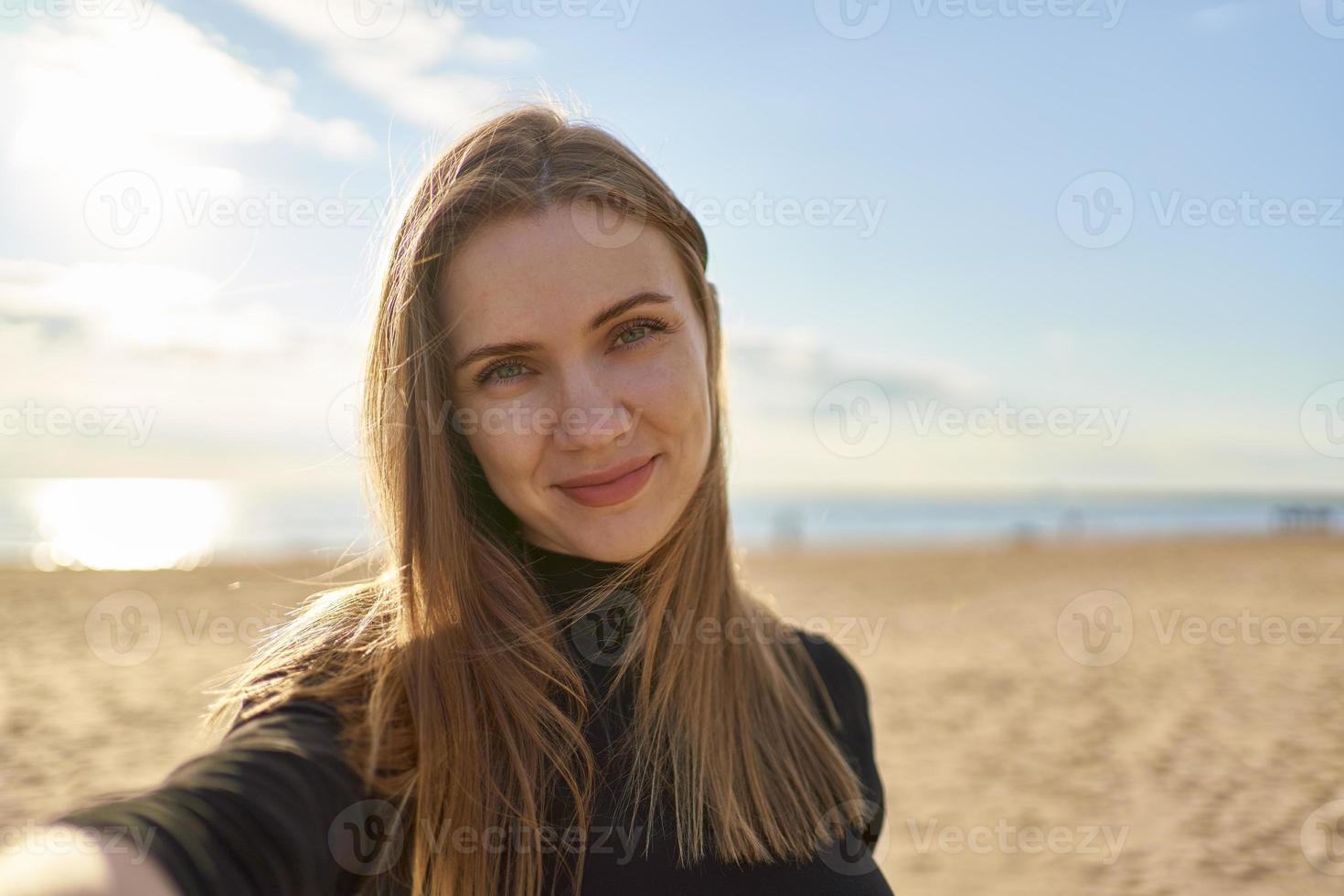 söt kvinna med långt hår tittar på kameran och ler i solig kväll i havet foto