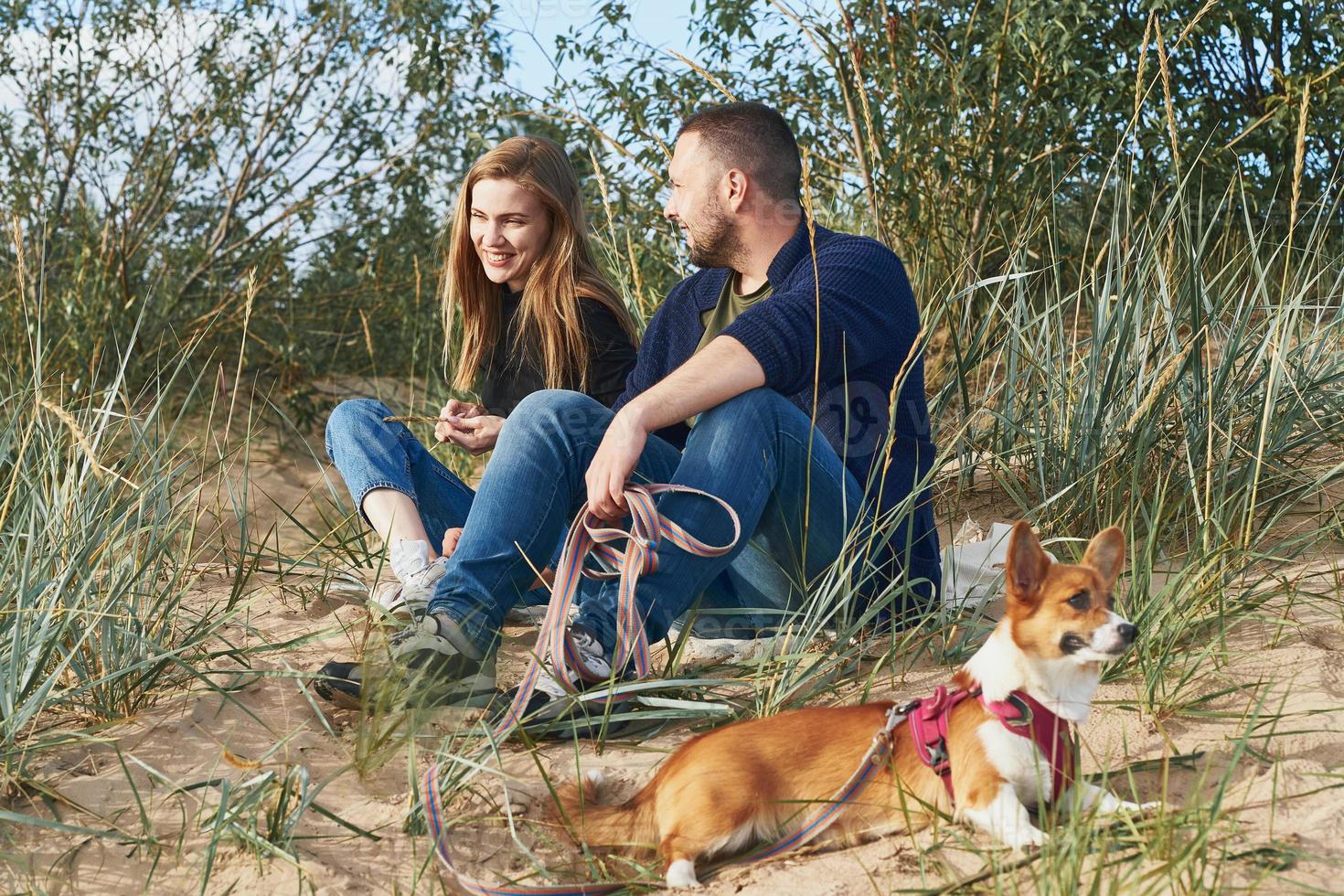 ungt lyckligt par av man och kvinna med corgihund som sitter i sanden. två personer med husdjur foto