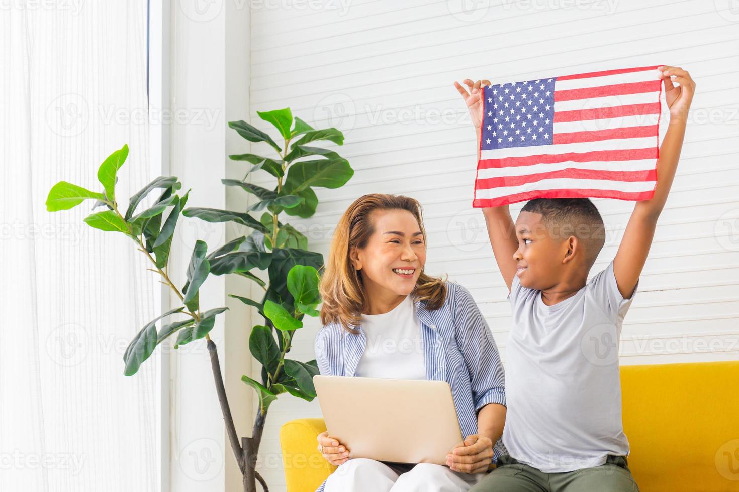 mormor och barnbarn leker glatt i vardagsrummet, kvinna med laptop och pojke håller USA flagga foto