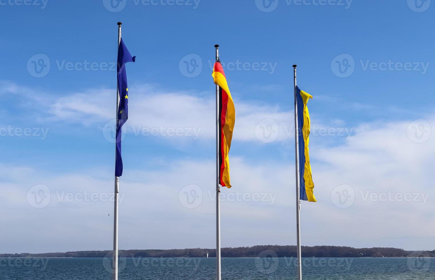 Ukrainas, Tysklands och Europeiska unionens flaggor flyger sida vid sida mot blå himmel. foto