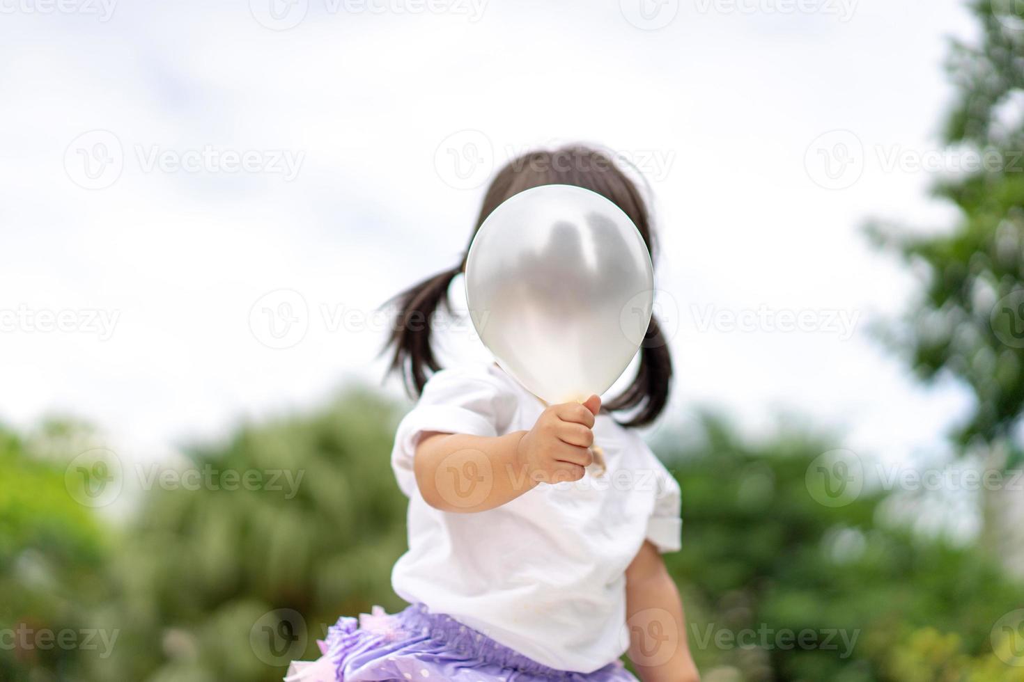 liten flicka håller den vita ballongen uppe på framsidan av hennes ansikte i trädgården. foto