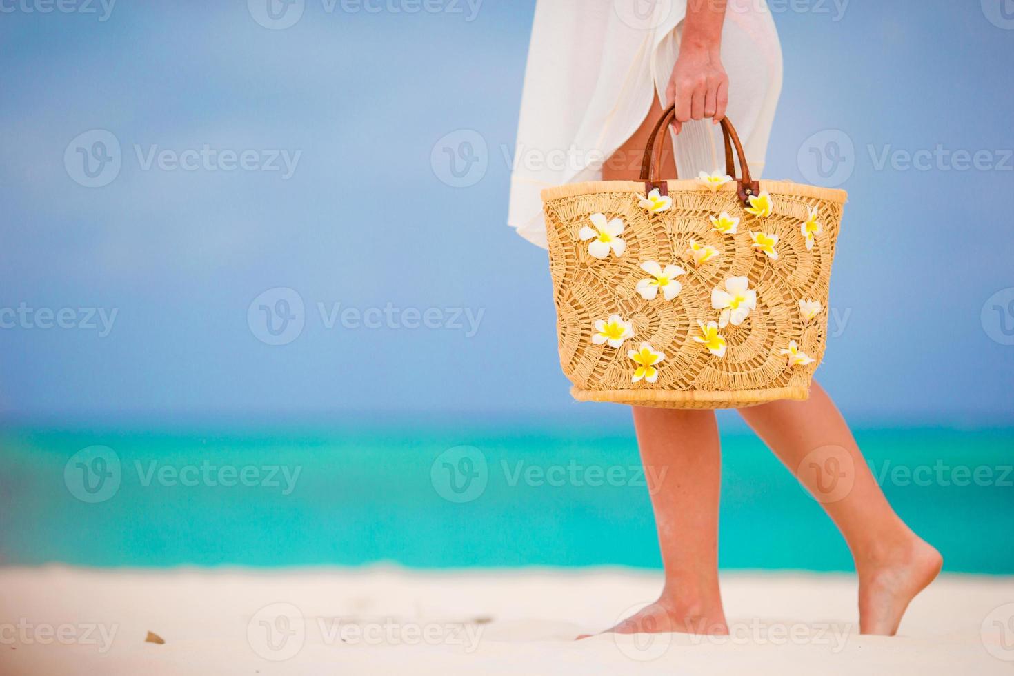 närbild vacker väska med frangipani blommor och solglasögon på vit strand i kvinnliga händer foto