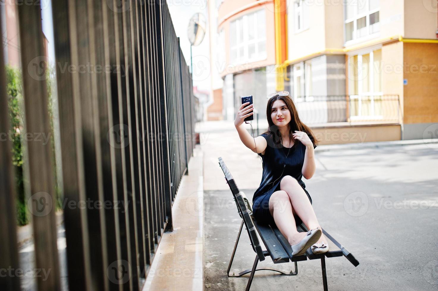 brunett flicka på svart klänning, solglasögon sitter på bänken, lyssnar på musik från hörlurar telefon och poserar på gatan i staden. foto