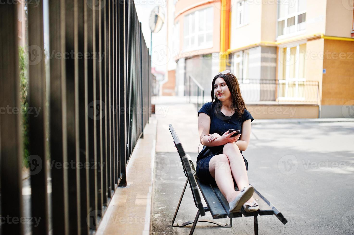 brunett flicka på svart klänning, solglasögon sitter på bänken, lyssnar på musik från hörlurar telefon och poserar på gatan i staden. foto