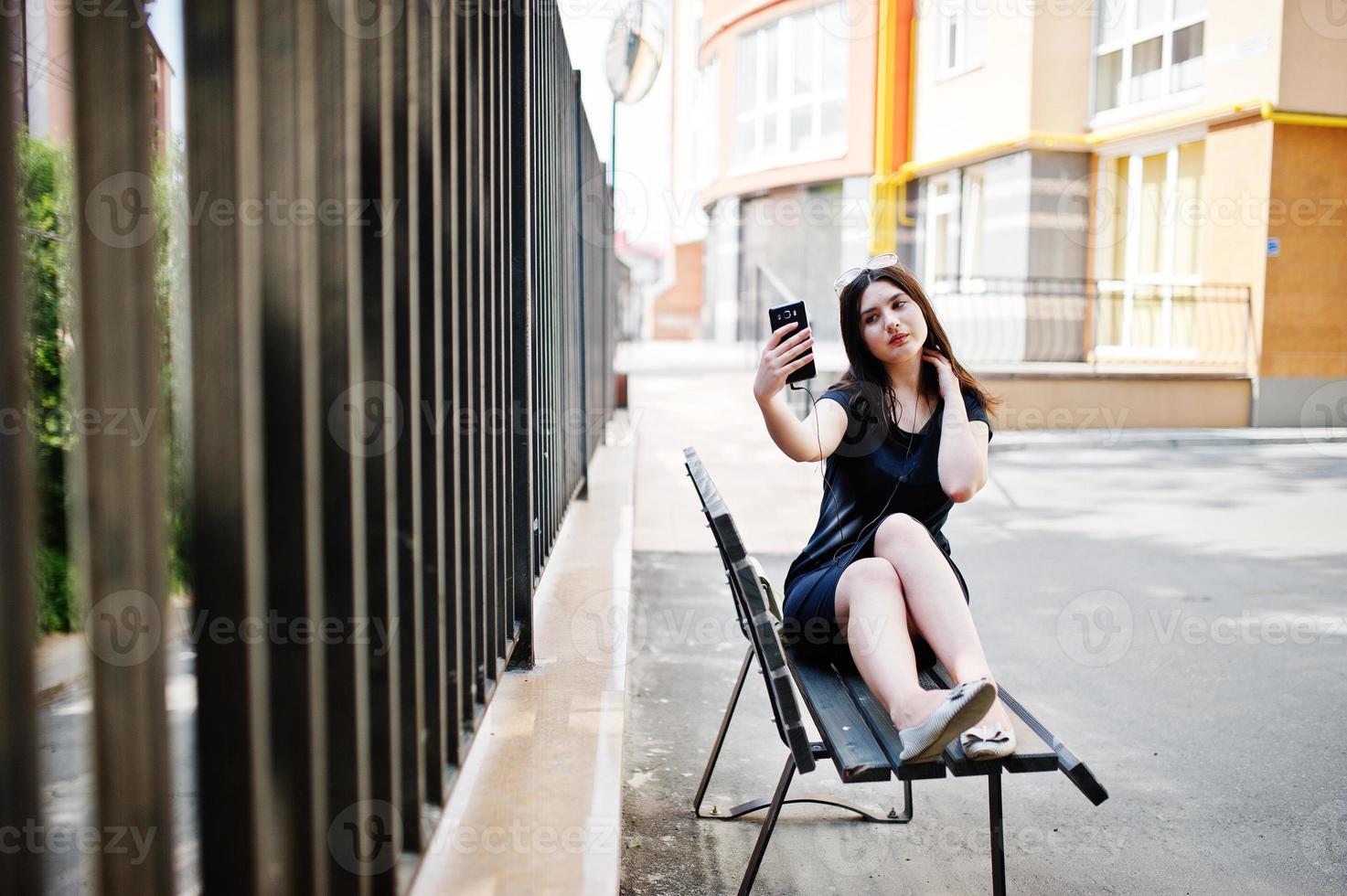 brunett flicka på svart klänning, solglasögon sitter på bänken, lyssnar på musik från hörlurar telefon och poserar på gatan i staden. foto