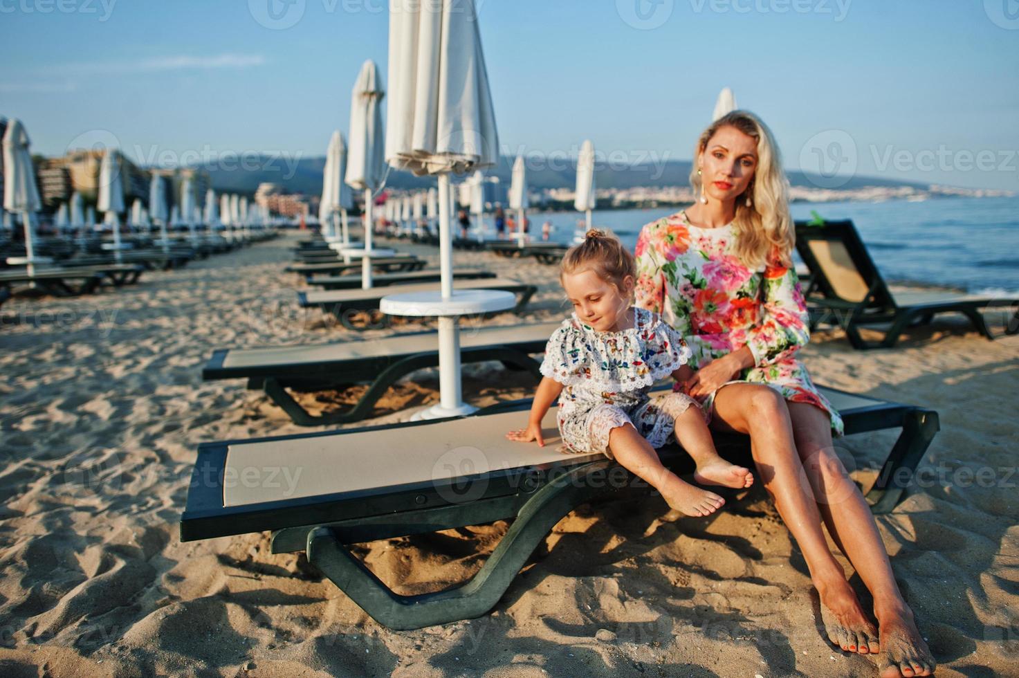 mamma och vackra dotter ha kul på stranden, sitter på solsäng. porträtt av glad kvinna med söt liten flicka på semester. foto