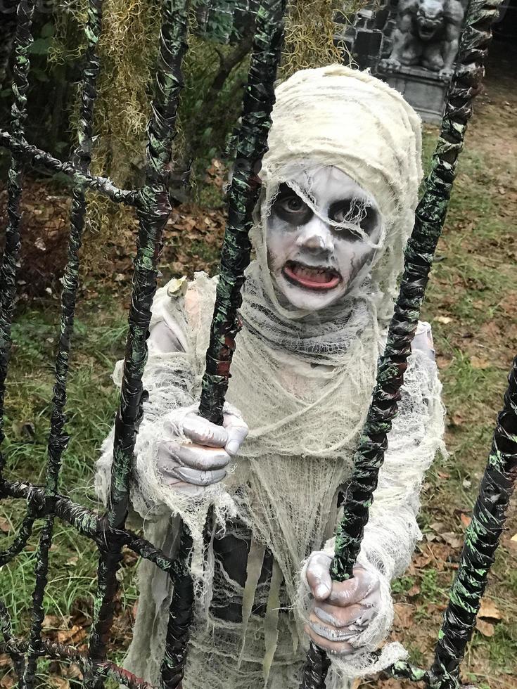 porträtt av ung pojke i kostym klädd som en halloween, cosplay av läskig mamma vid kyrkogårdens staket foto