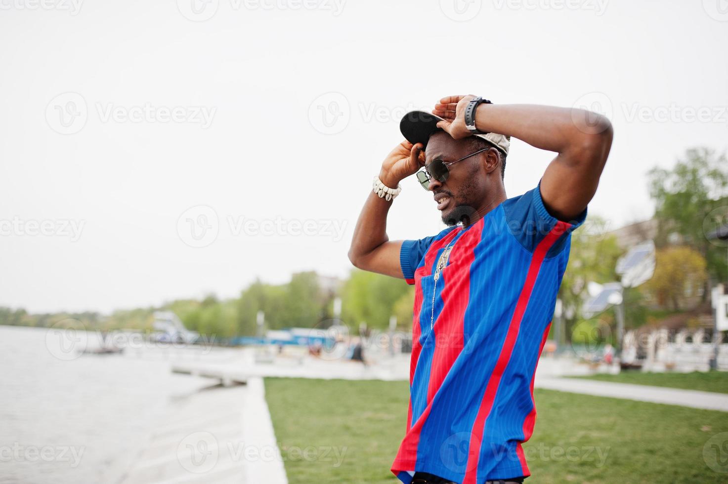 snygg afrikansk amerikansk pojke på keps, fotbollströja och solglasögon stannar mot lake beach. svart sport man porträtt. foto