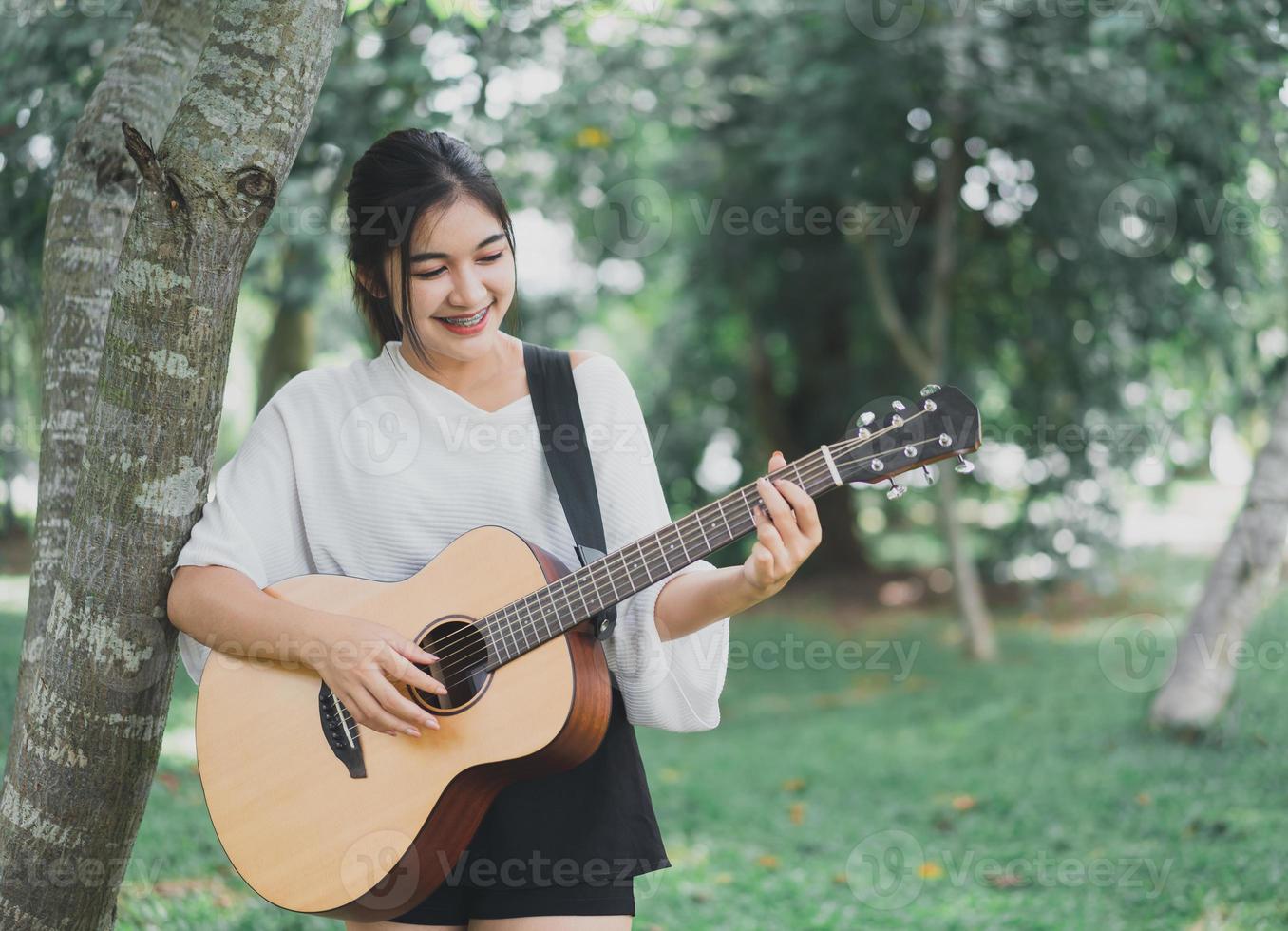 asiatisk ung kvinna spelar gitarr och sjunger musik i parken, asiatisk kvinna spelar gitarr i trädgården foto