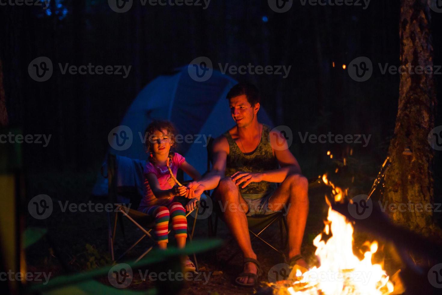 pappa och dotter sitter på natten vid brasan i det fria på sommaren i naturen. familjecamping, samlingar kring lägerelden. fars dag, grillning. campinglykta och tält foto
