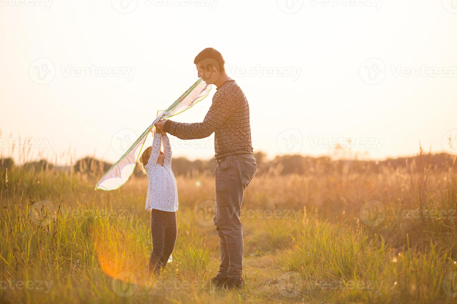 pappa hjälper sin dotter att flyga en drake på ett fält på sommaren vid solnedgången. familjeunderhållning utomhus, fars dag, barnens dag. landsbygd, stöd, ömsesidig hjälp. orange ljus från solen foto