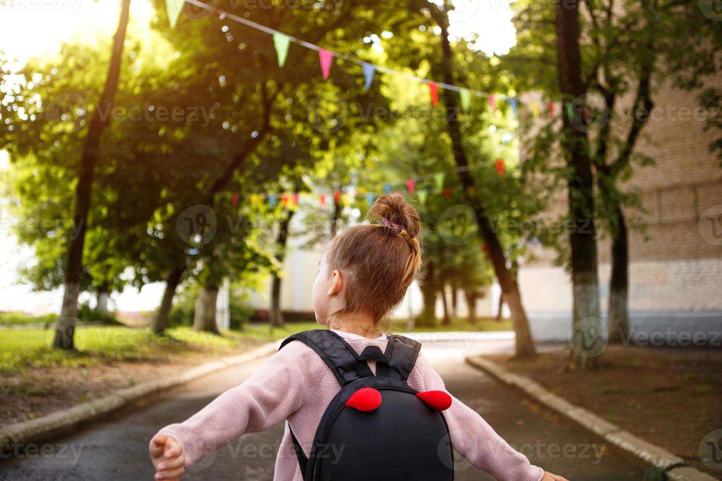 en liten flicka av kaukasiska i en skoluniform med en ryggsäck tittar på vägen på skolgården. koncept tillbaka till skolan. grundskola, utveckla verksamhet för förskolebarn. utrymme för text foto