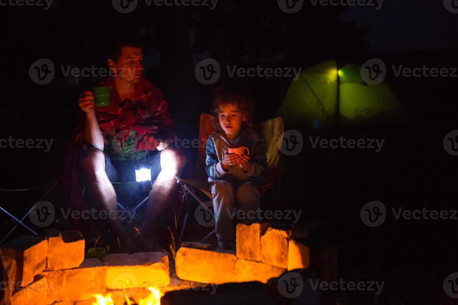 pappa och dotter sitter på natten vid brasan i det fria på sommaren i naturen. familjecamping, samlingar kring lägerelden. fars dag, grillning. campinglykta och tält foto