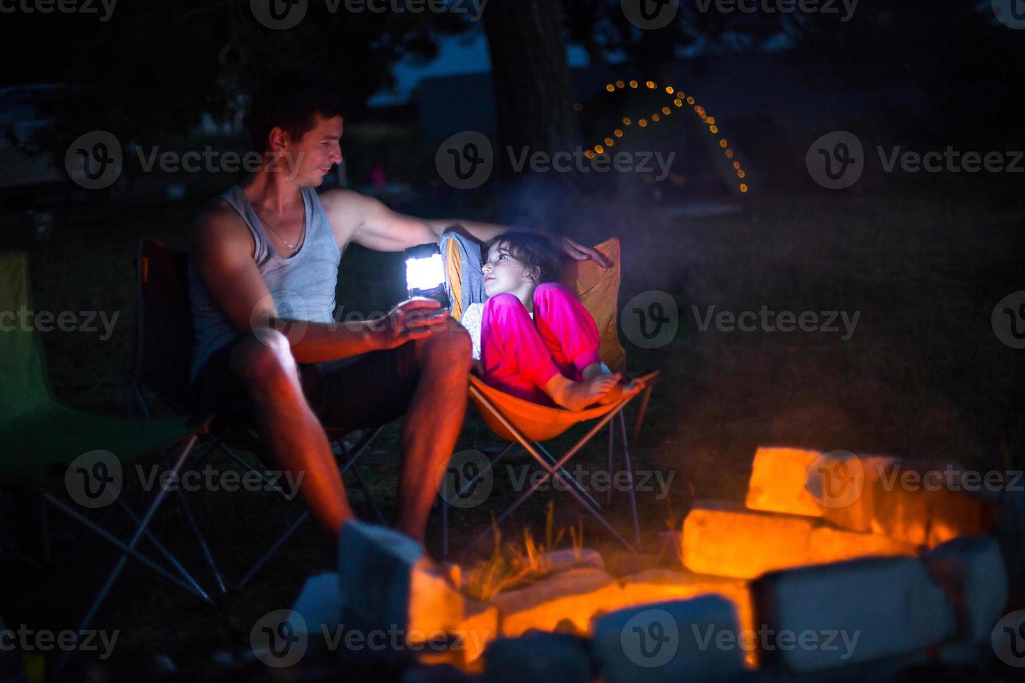 pappa och dotter sitter på natten vid brasan i det fria på sommaren i naturen. familjecamping, samlingar kring lägerelden. fars dag, grillning. campinglykta och tält foto