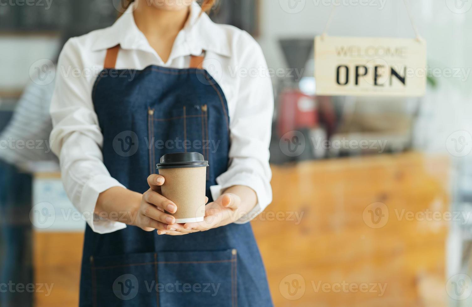 porträtt av glad kvinna som står vid dörröppningen till hennes butik. glad mogen servitris som väntar på kunder på kaféet. framgångsrik småföretagare i casual klädd i blått förkläde stående vid ingången foto