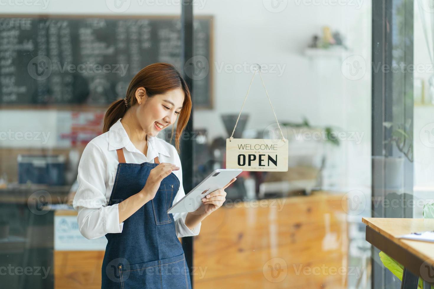 porträtt av glad kvinna som står vid dörröppningen till hennes butik. glad mogen servitris som väntar på kunder på kaféet. framgångsrik småföretagare i casual klädd i blått förkläde stående vid ingången foto