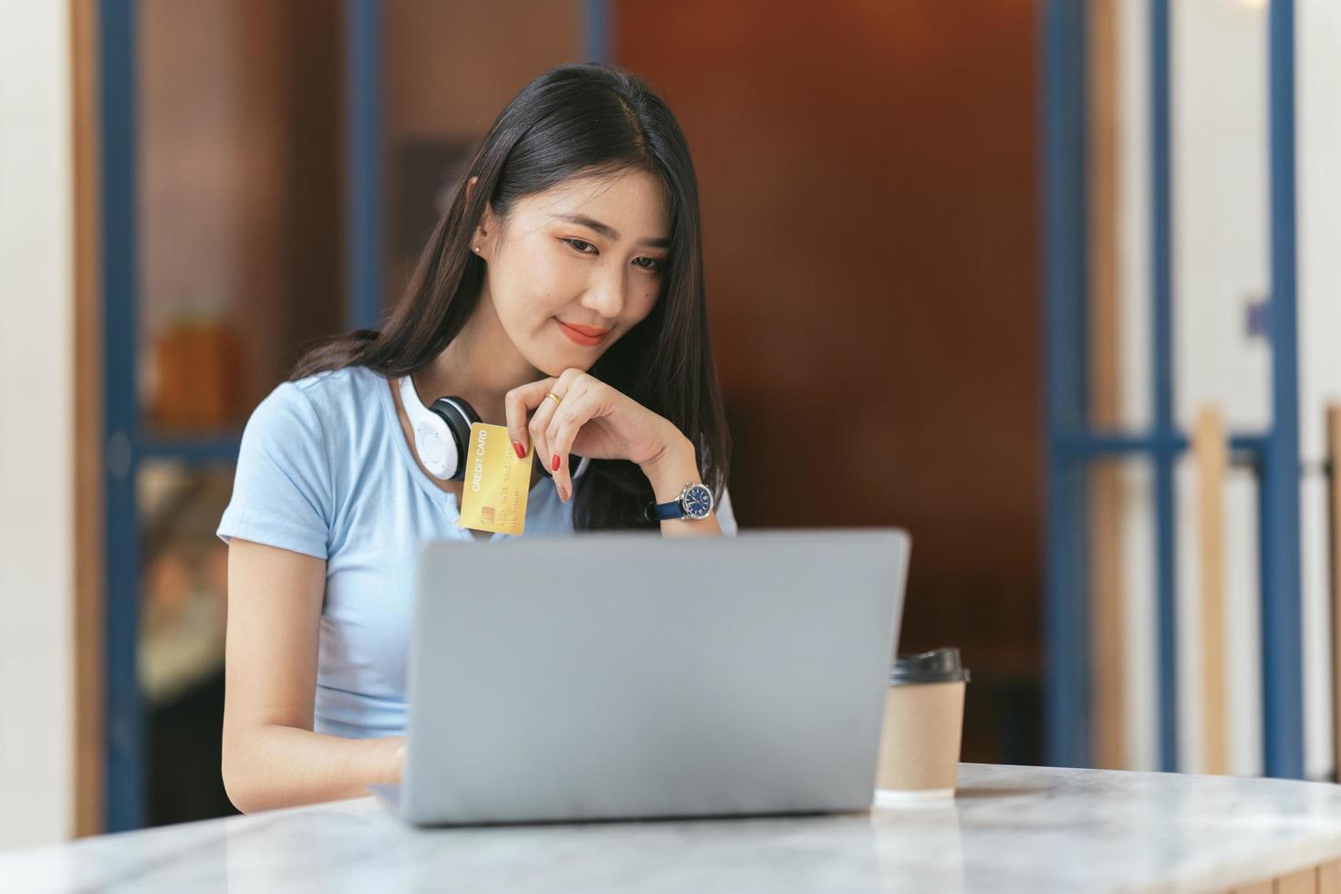 online bankkoncept. porträtt av glad ung asiatisk kvinna med bärbar dator och kreditkort sitter på café, leende asiatiska kvinnor njuter av att göra betalningar hemifrån. foto