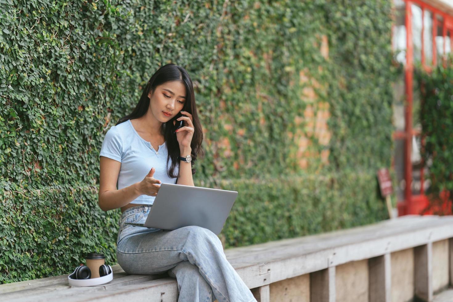 porträtt av vacker asiatisk kvinna som sitter utomhus under sommaren, använder smart trådlös teknik dator bärbar dator och smartphone, avkopplande kaffepaus på café restaurang. foto