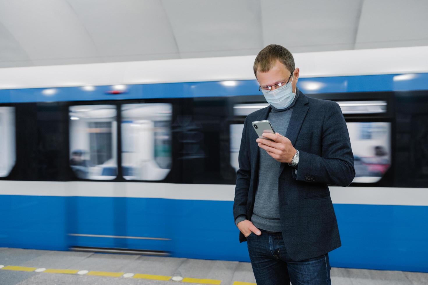 manliga passagerare bär ansiktsmask poserar vid plattformen, väntar på tåget, pendlar med tunnelbanan, koncentrerad i smartphone-enhet, läser nyheter online. virusmedvetenhet på offentlig plats. coronavirus utbrott foto