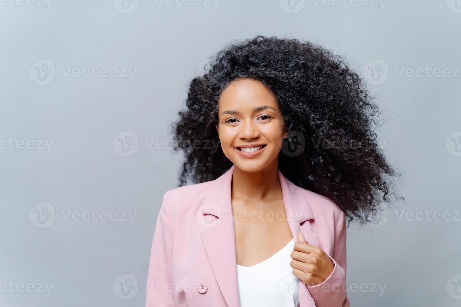 positiv lockig mörkhyad kvinna med självlysande hår, bär formell lila jacka, poserar mot grå bakgrund med tomt utrymme, glad att träffa affärspartners. känslor. glad regissör foto