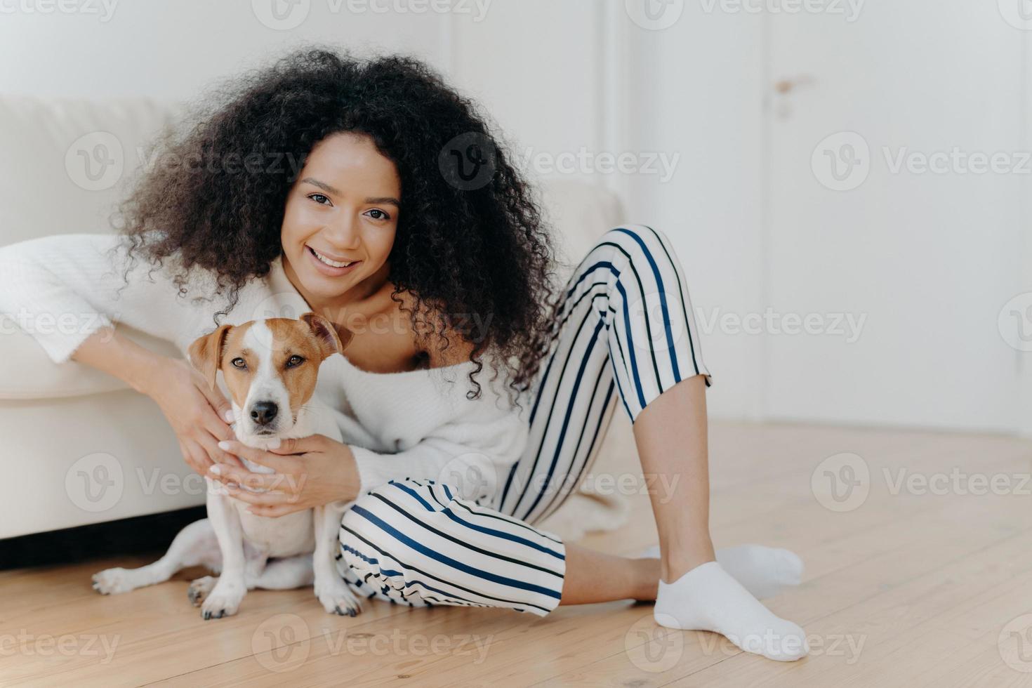inomhusbild av nöjd lockig kvinnlig modell poserar för foto med jack russell terrierhund, omfamnar husdjur ömt, sitter på golvet med älskade djur nära en bekväm soffa, umgås tillsammans