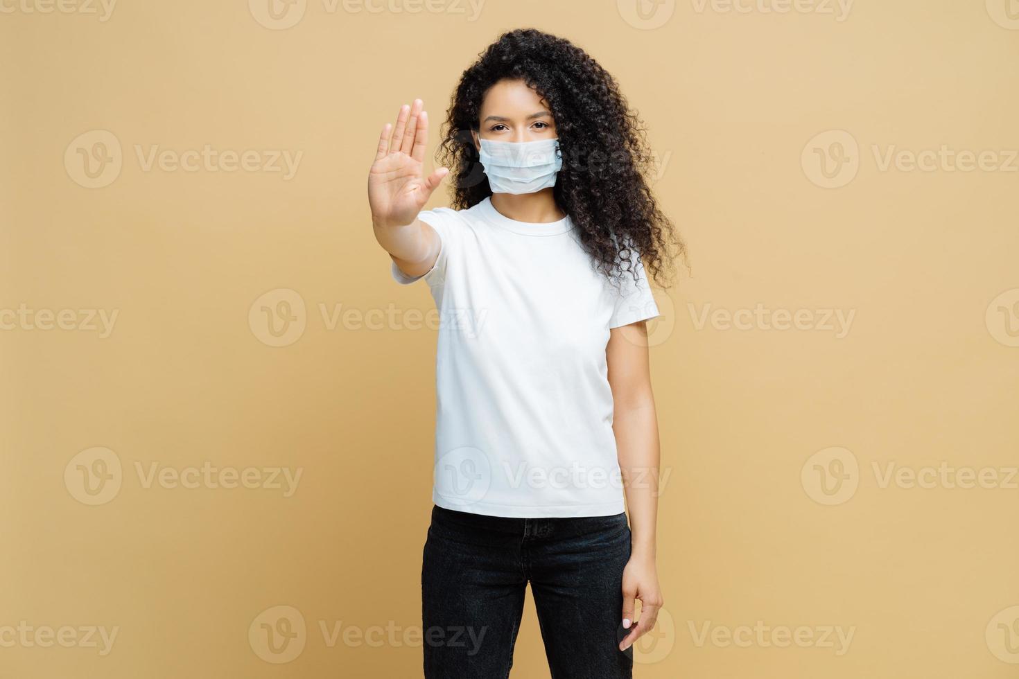 säga nej till att sprida sjukdomar. seriös afroamerikansk kvinna gör en stoppgest, bär medicinsk mask, förhindrar virus covid-19, står inomhus, isolerad. stoppa infektionssjukdomar omedelbart foto