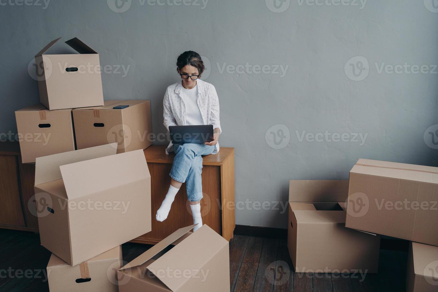 spansk affärskvinna hyr lägenhet. glad tjej sitter bland lådor och arbetar på laptop. foto