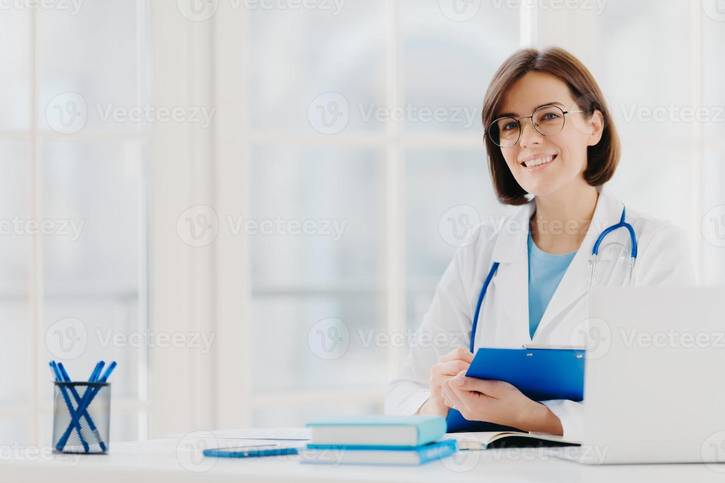 kvinnlig läkare skriver ut recept på specialformulär, arbetar på privat klinik, bär vit sjukvårdsrock, redo att träffa patienter, poserar på arbetsplatsen. leende läkare eller medicinsk arbetare håller urklipp foto