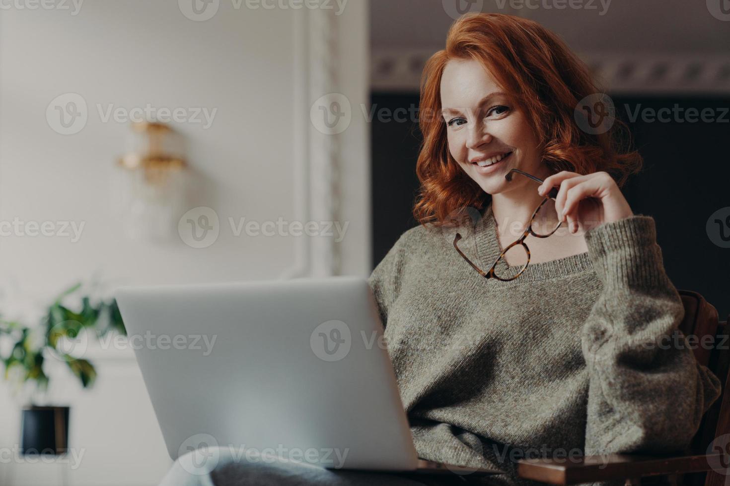 självsäker glad rödhårig kvinna håller glasögon för synkorrigering har fjärrjobb upptagen med onlineprojekt bär varm tröja, sitter framför bärbar dator, planerar effektivt arbete foto