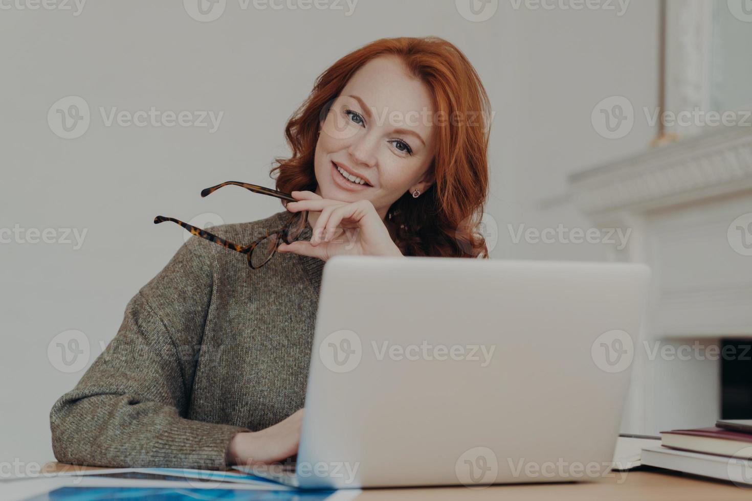 foto av glad rödhårig kvinnlig student gör forskning för läxor, sitter på coworking utrymme i studierummet, arbetar på bärbar dator, använder internet. frilansande webbdesigner skapar design för klient